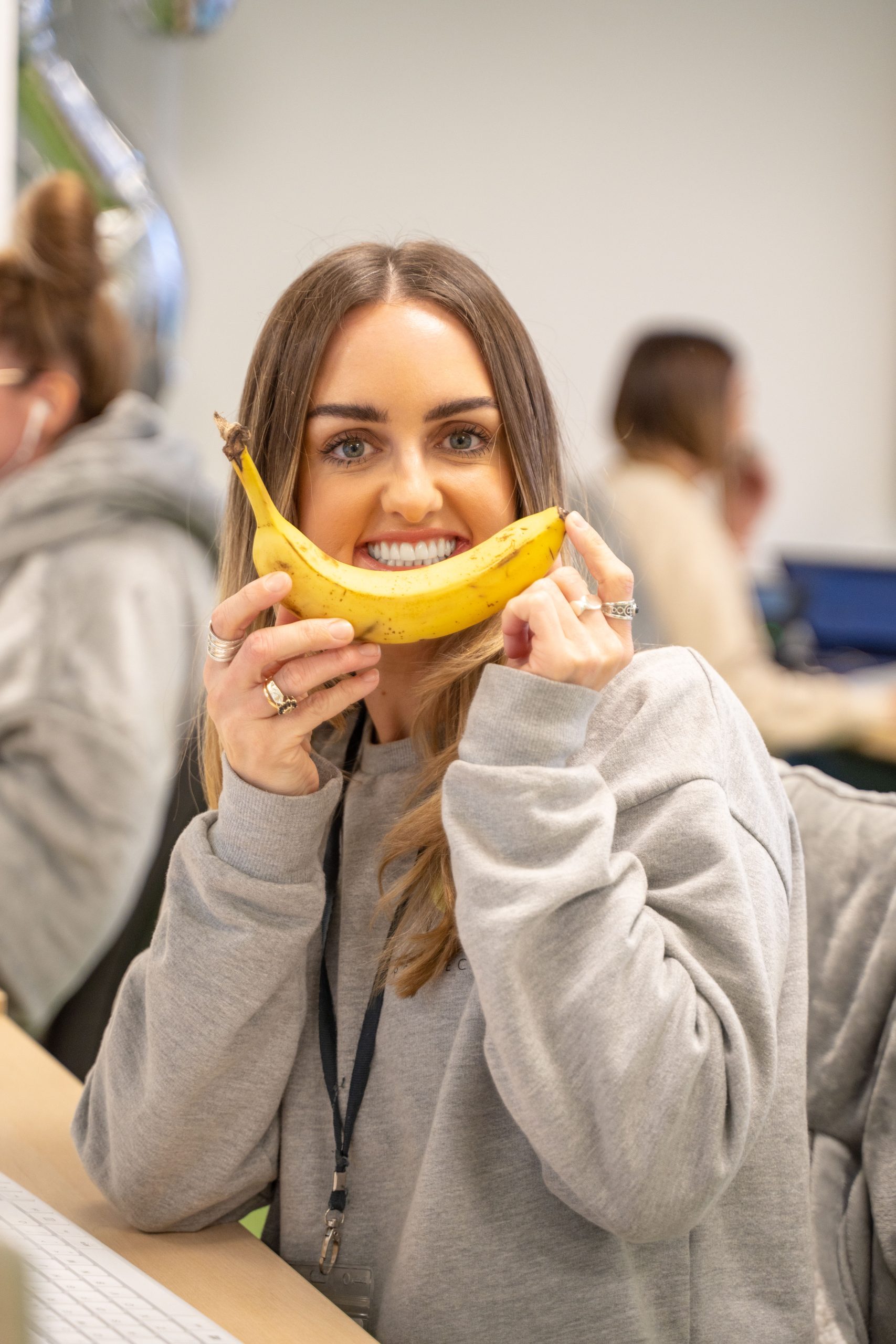Emily - sat at her desk with a smile on her face as she is holding a banana in front of her face, replicating a smile.